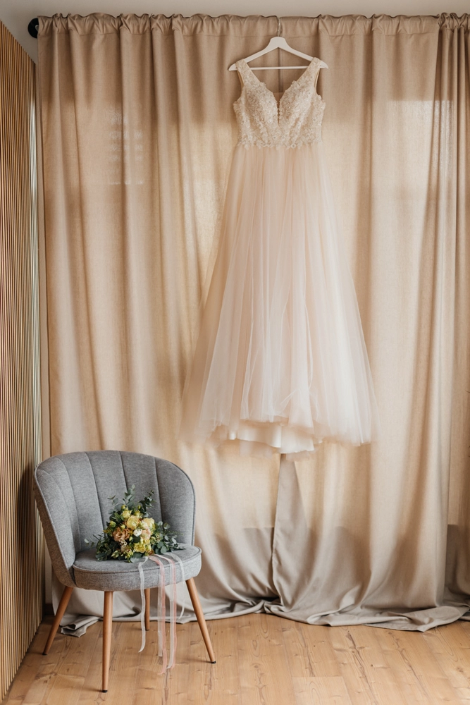 Ein elegantes Brautkleid hängt stimmungsvoll vor einem hellen Vorhang, bereit für das Getting Ready.