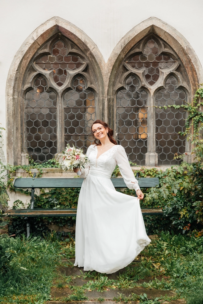 Braut im langen weißen Kleid vor historischen Spitzbogenfenstern