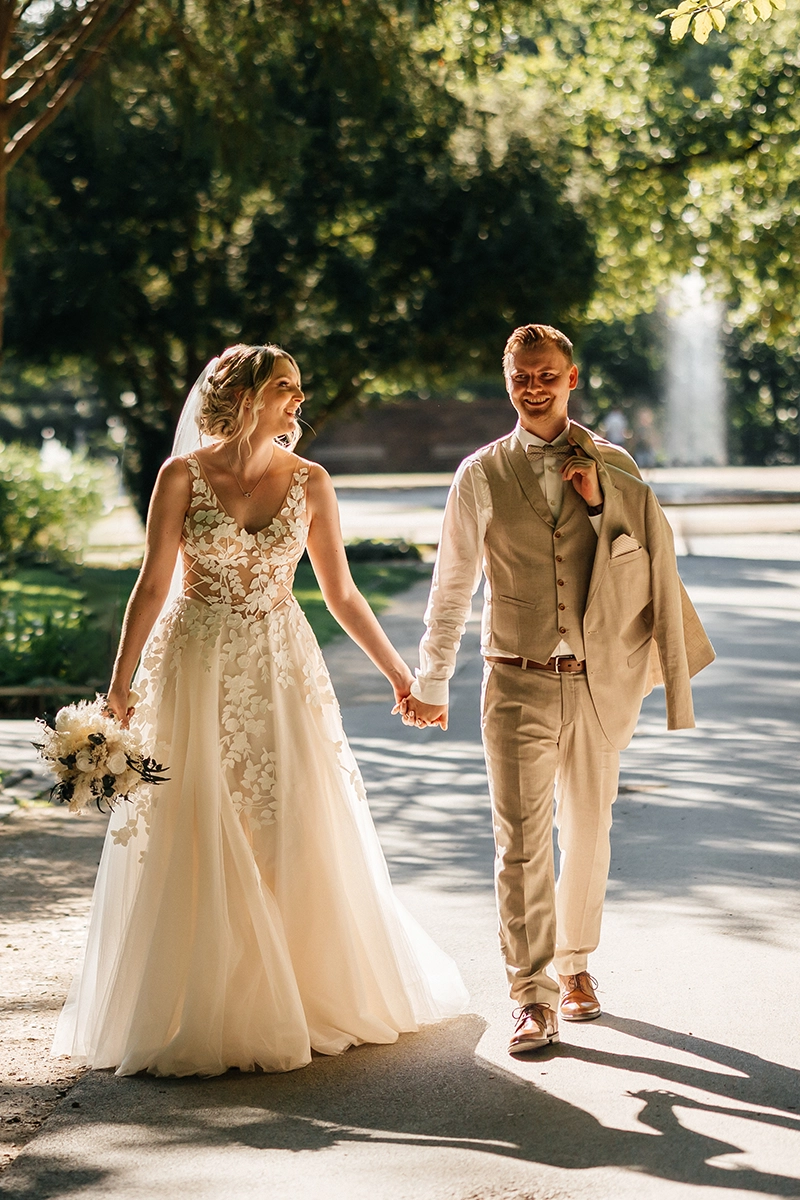 Brautpaar läuft Hand in Hand und lachend auf die Kamera zu, entspannte Bewegung im Park.