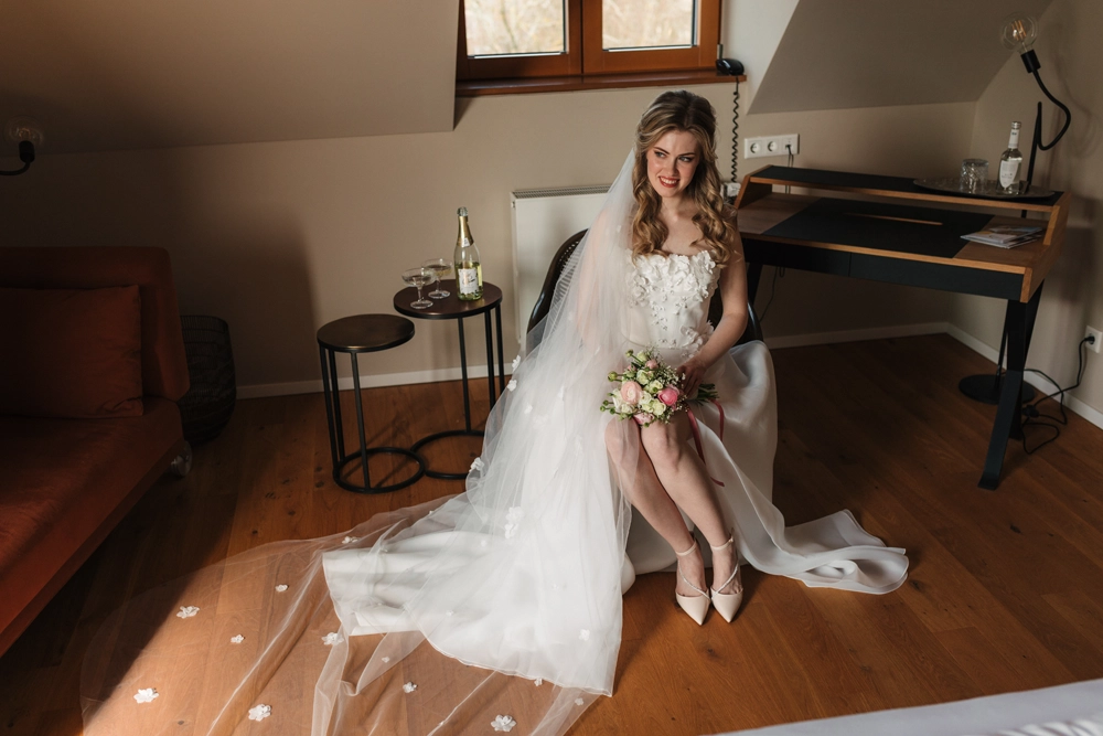 Die junge Braut mit lockigem, blondem Haar trägt ein Kleid mit Blumenapplikationen. Ihr Blumenstrauß aus Rosa- und Weißtönen rundet den romantischen Look ab.