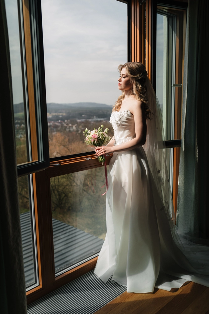 Stilvolles Brautporträt: Eine blonde Frau im weißen Kleid mit floralen Details hält einen Strauß aus weißen und rosafarbenen Rosen und schaut aus dem Fenster ihres Hotelzimmer