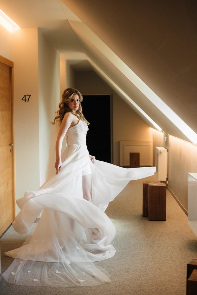 Braut mit langen, welligen blonden Haaren steht in einem Hotelflur vor einer Zimmertür mit der Nummer 47. Sie trägt ein weisses Hochzeitskleid mit fliessendem Stoff, der beim Drehen in Bewegung ist. Das Kleid gibt den Blick auf ihre Beine und weisse Strümpfe frei. Die Szenerie ist modern und warm beleuchtet.