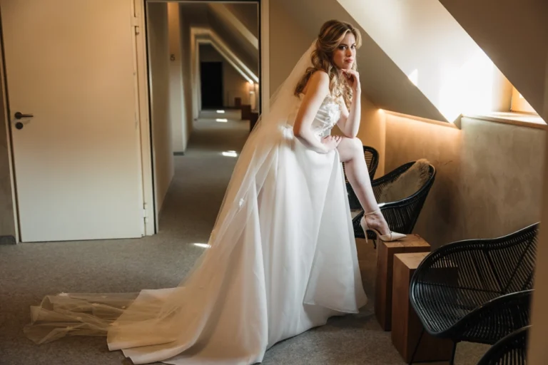 Verspielte Brautszene im Hotelflur: Die junge Frau mit blondem Haar trägt ein weißes Kleid und zeigt ihr Bein mit Brautschuh