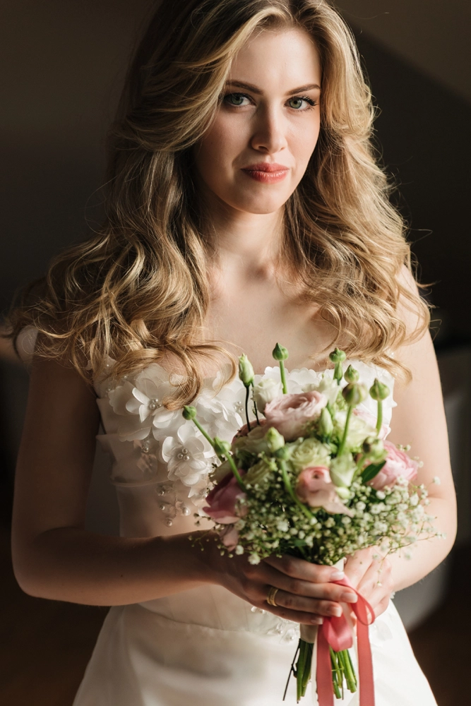 Porträt einer jungen Braut mit langen, blonden Locken. Sie trägt ein weißes Kleid mit Blumen-Details und hält einen zarten Strauß aus rosa und weißen Rosen. Der Hintergrund ist dunkel und unscharf.