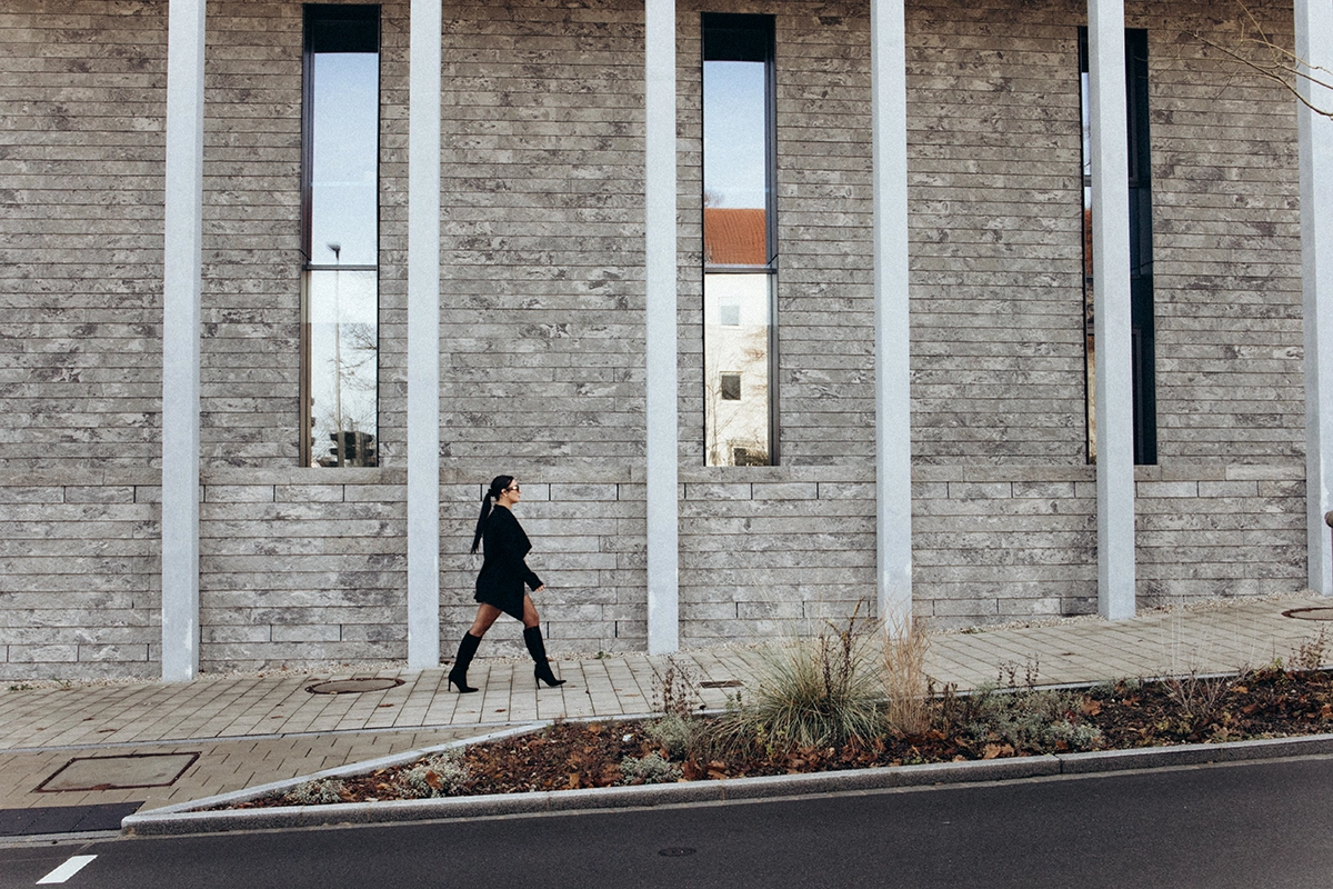 Dynamisches Businessportrait im Editorial-Stil: Frau läuft selbstbewusst vor moderner Beton-Architektur, Business-Casual Look mit Mantel.
