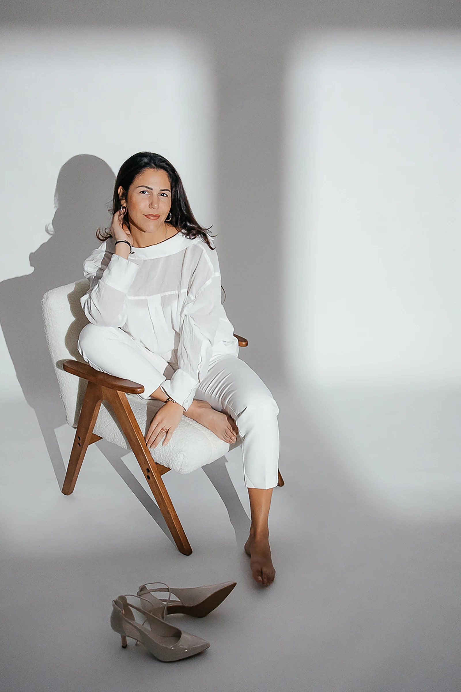 Minimalistisches Personal Branding Foto im Studio: Frau in weißem Outfit sitzt entspannt auf einem Stuhl, High-Fashion Pose ohne Schuhe.