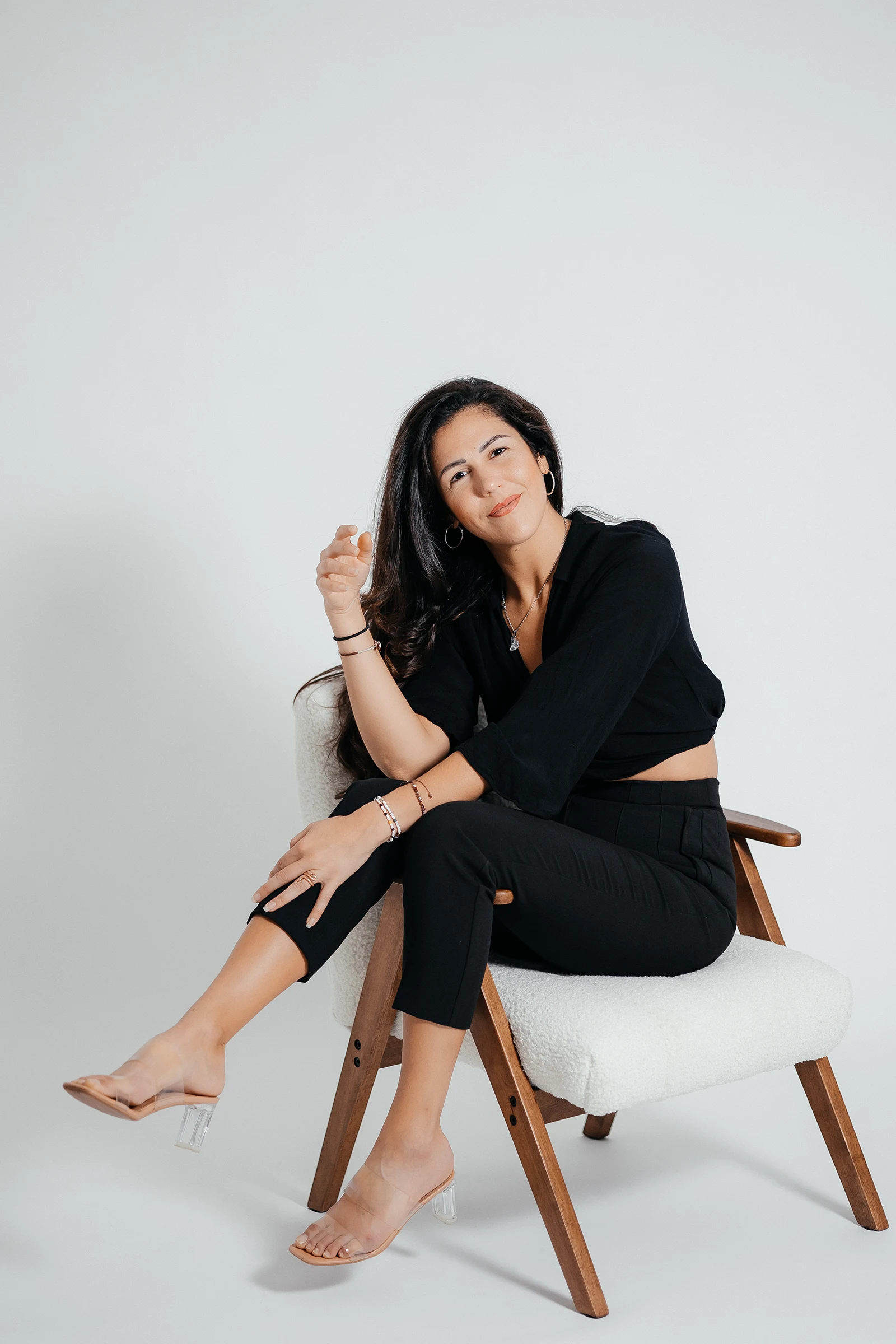 Minimalistisches Personal Branding Foto im Studio: Frau in schwarzem Zweiteiler sitzt entspannt und barfuß auf einem Designer-Holzsessel, lachend und nahbar.