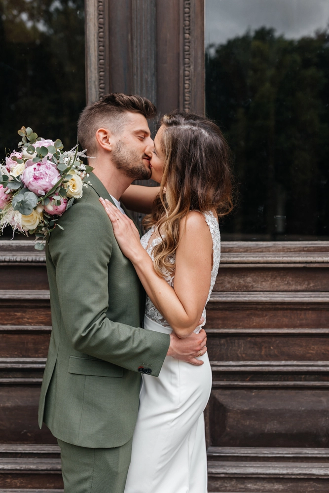 Inniger Kuss des Brautpaares vor dunklem Hintergrund, Braut hält eleganten Brautstrauß mit rosa und weißen Blumen.