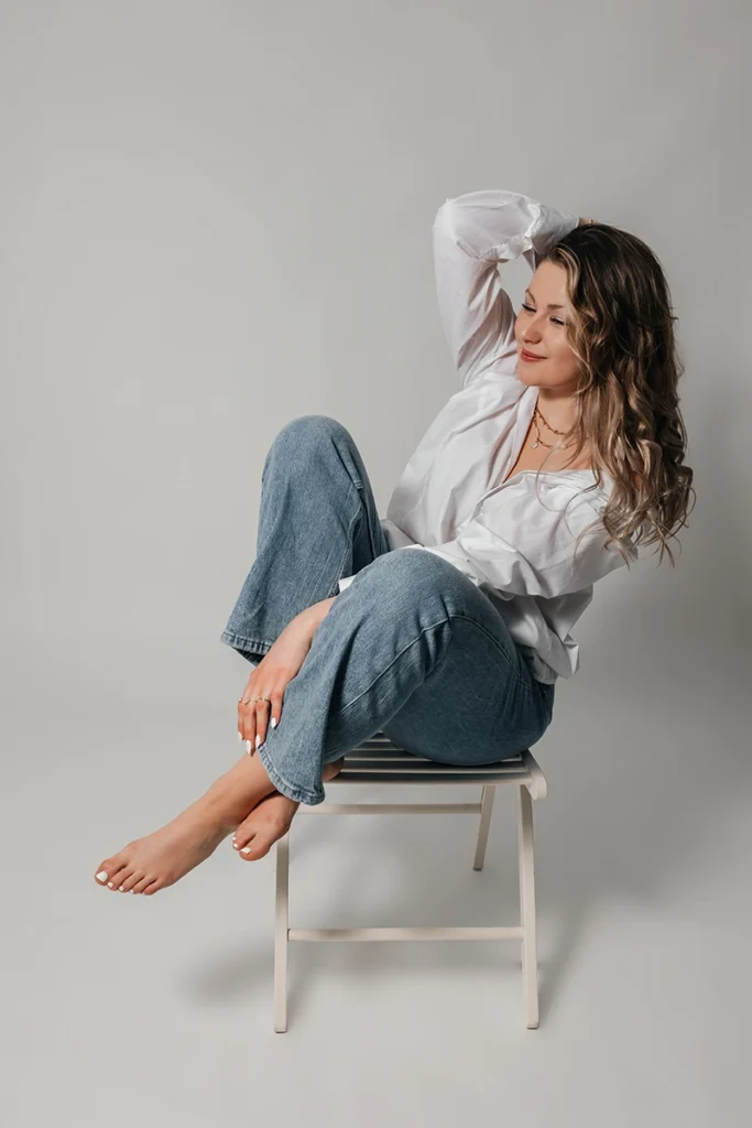Entspanntes Studio-Portrait in Burgthann: Frau in Jeans und weißem Hemd sitzt barfuß auf einem Stuhl und lacht natürlich.
