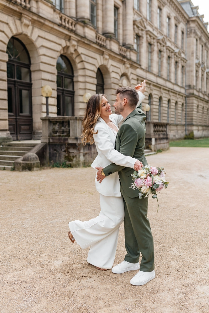 Braut im weißen Hosenanzug hebt spielerisch das Bein beim Kuss vor der Orangerie Erlangen, Bräutigam im grünen Anzug hält sie im Arm