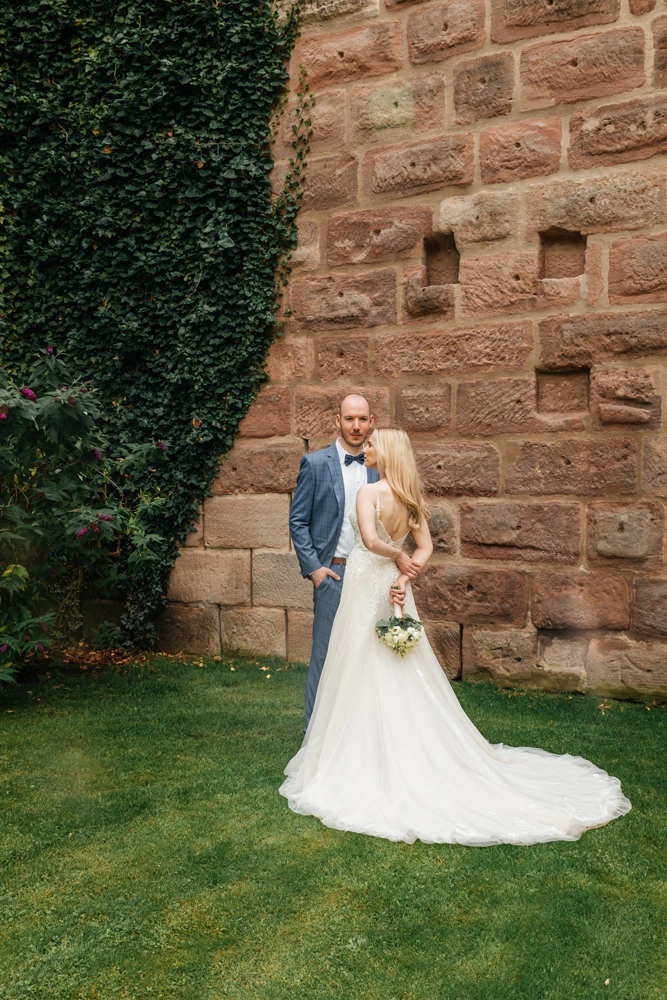 Brautpaar posiert vor einer massiven historischen Sandsteinmauer mit Efeu, minimalistisches Hochzeitsfoto.