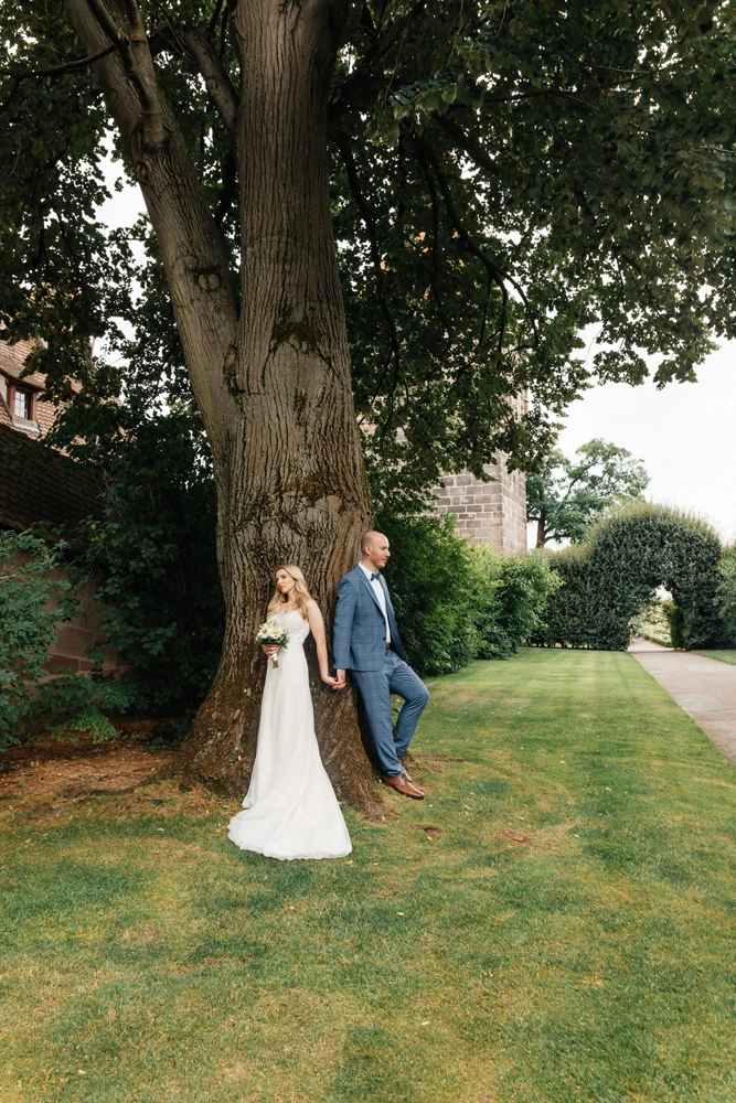 Brautpaar steht für einen intimen Moment unter einem großen Baum im Burggarten, Spiel mit Licht und Schatten.