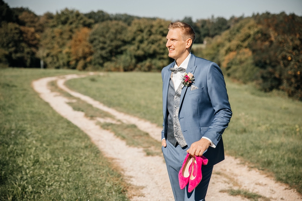 Bräutigam im blauen Anzug läuft auf Feldweg und trägt lässig die pinken High Heels der Braut.