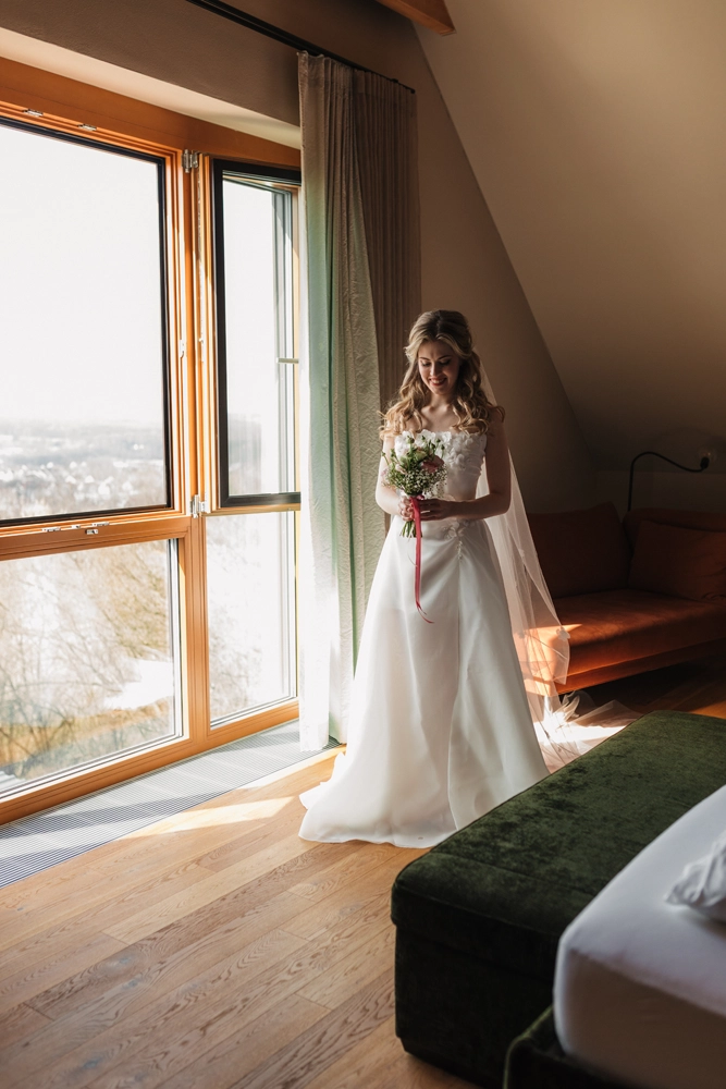ertig gestylte Braut im Hochzeitskleid steht elegant am Fenster.