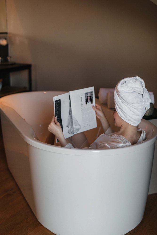 Braut mit Handtuch-Turban sitzt entspannt in einer weißen Badewanne und vertieft sich in ein Magazin.