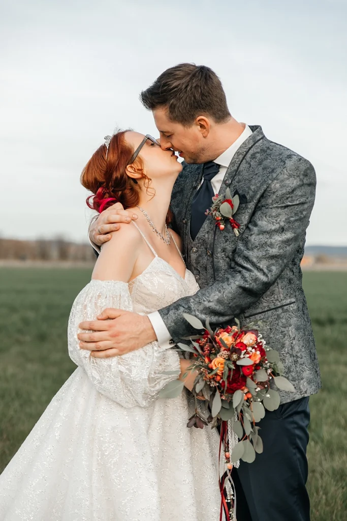 Romantischer Kuss im Feld: Braut mit Brille und trendigem Brautkleid mit großen Puffärmeln (Statement Sleeves). Der Bräutigam trägt einen gemusterten Anzug. Herbstlicher Brautstrauß in Rot- und Orangetönen