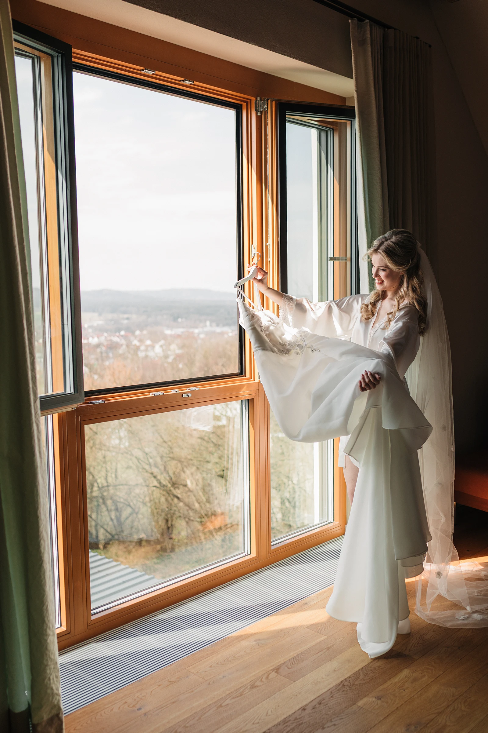 Braut steht am großen Fenster und öffnet die Vorhänge, schönes Morgenlicht.