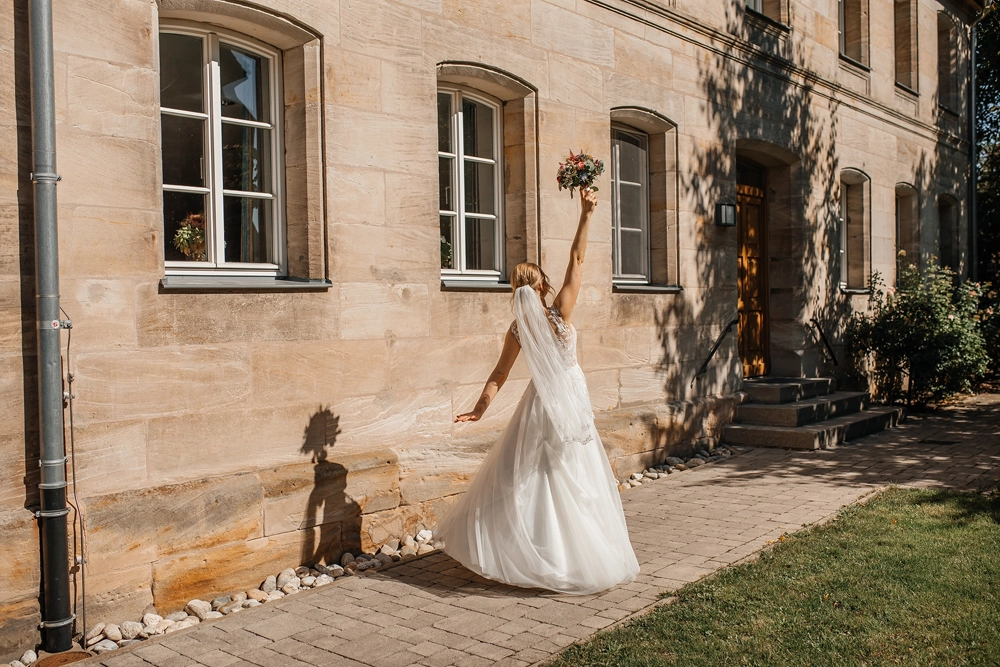 Braut wirft lachend den Arm in die Luft vor der hellen Kirchenmauer.