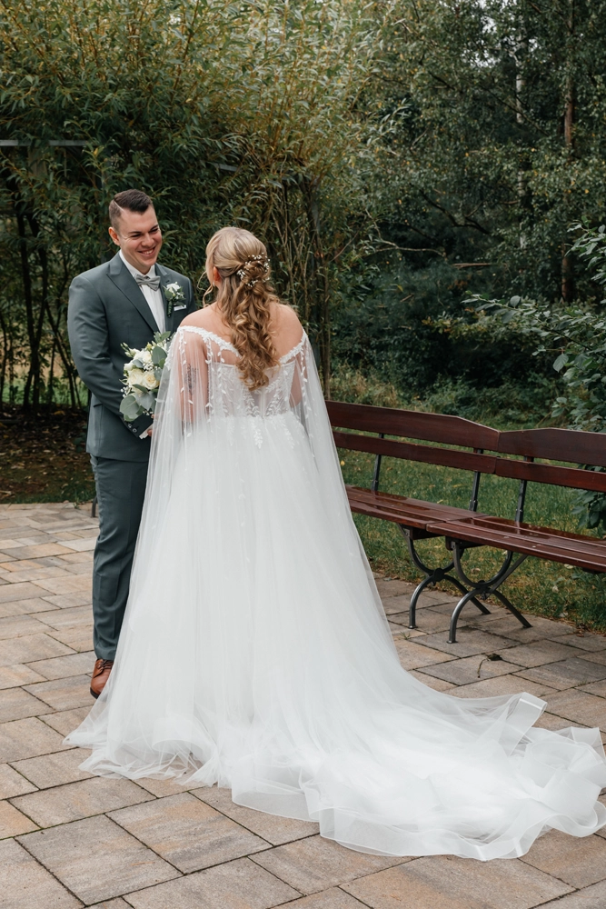 Rückenansicht des Brautkleids mit bestickten Cape-Ärmeln und Schleier-Effekt im Grünen.