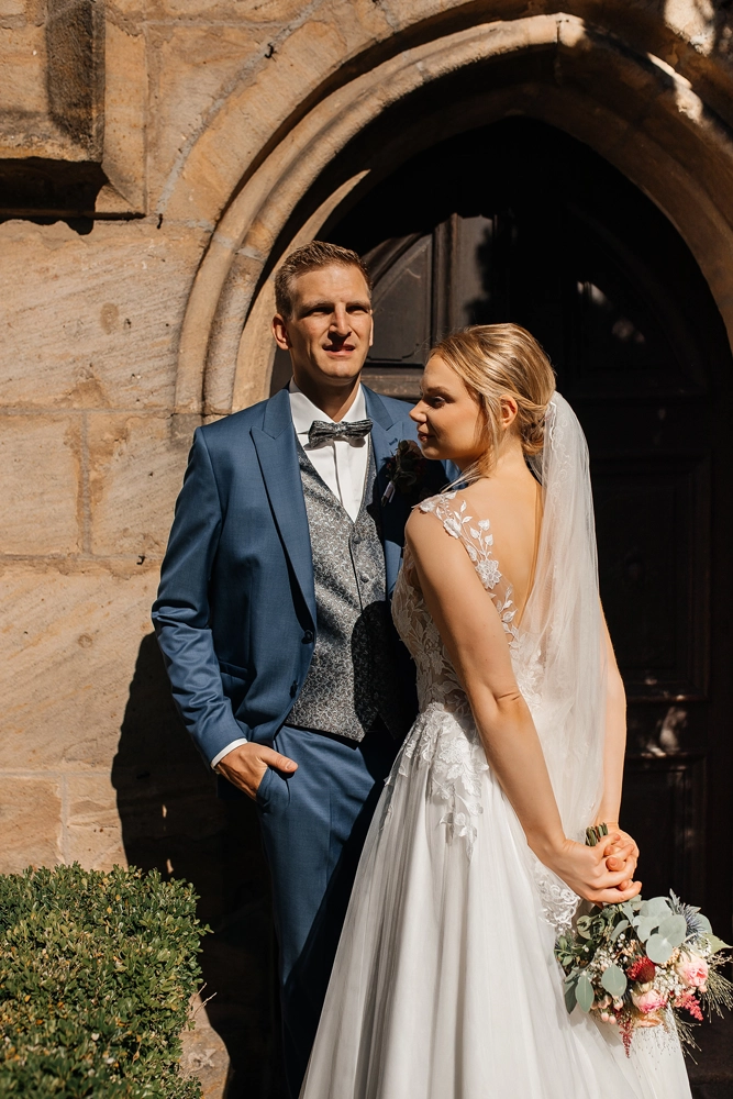 Braut und Bräutigam stehen vor dem massiven dunklen Holzportal im Sonnenlicht.