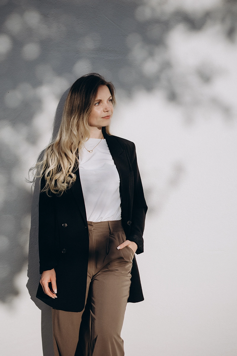 Modernes Business-Outfit Inspiration: Frau mit schwarzem Blazer, weißem Shirt und beiger Hose steht draußen, Blick selbstbewusst zur Seite.