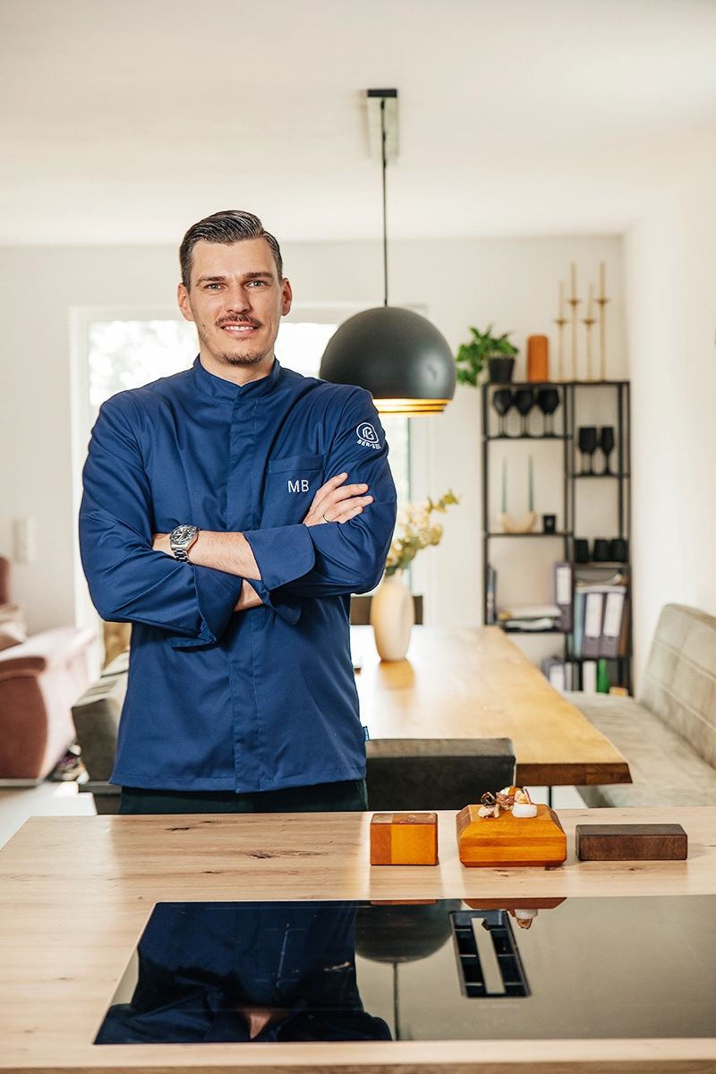 Modernes Business Portrait von Sternekoch Marcus Bunzel in blauer Kochjacke, stehend in seiner Küche.