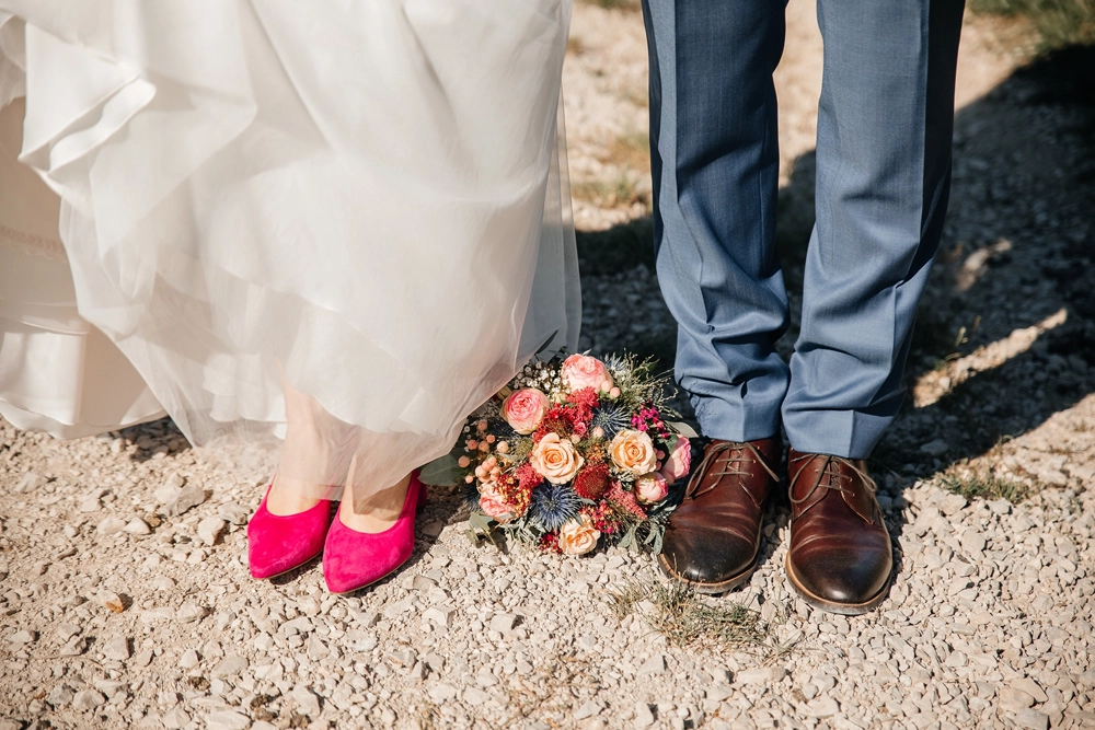 Detailaufnahme der Füße: Braut in knallpinken Pumps neben dem Bräutigam auf Kiesweg