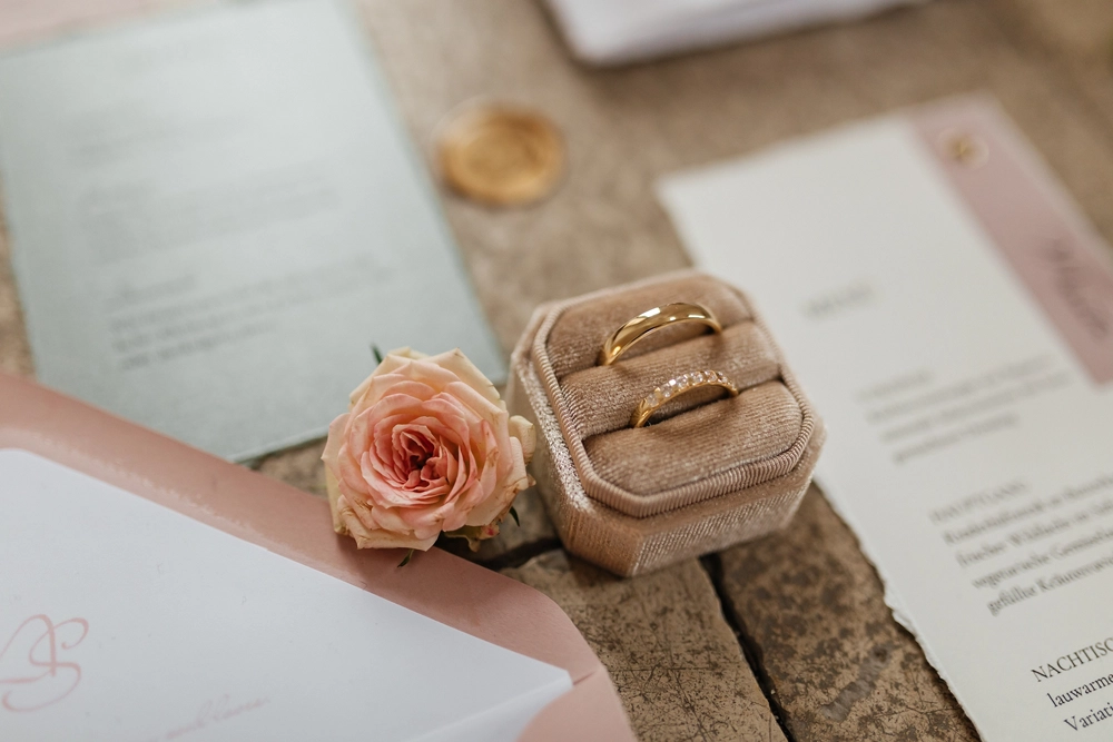Detailaufnahme goldener Eheringe in einer beigen Samtbox, dekoriert mit einer Rose.