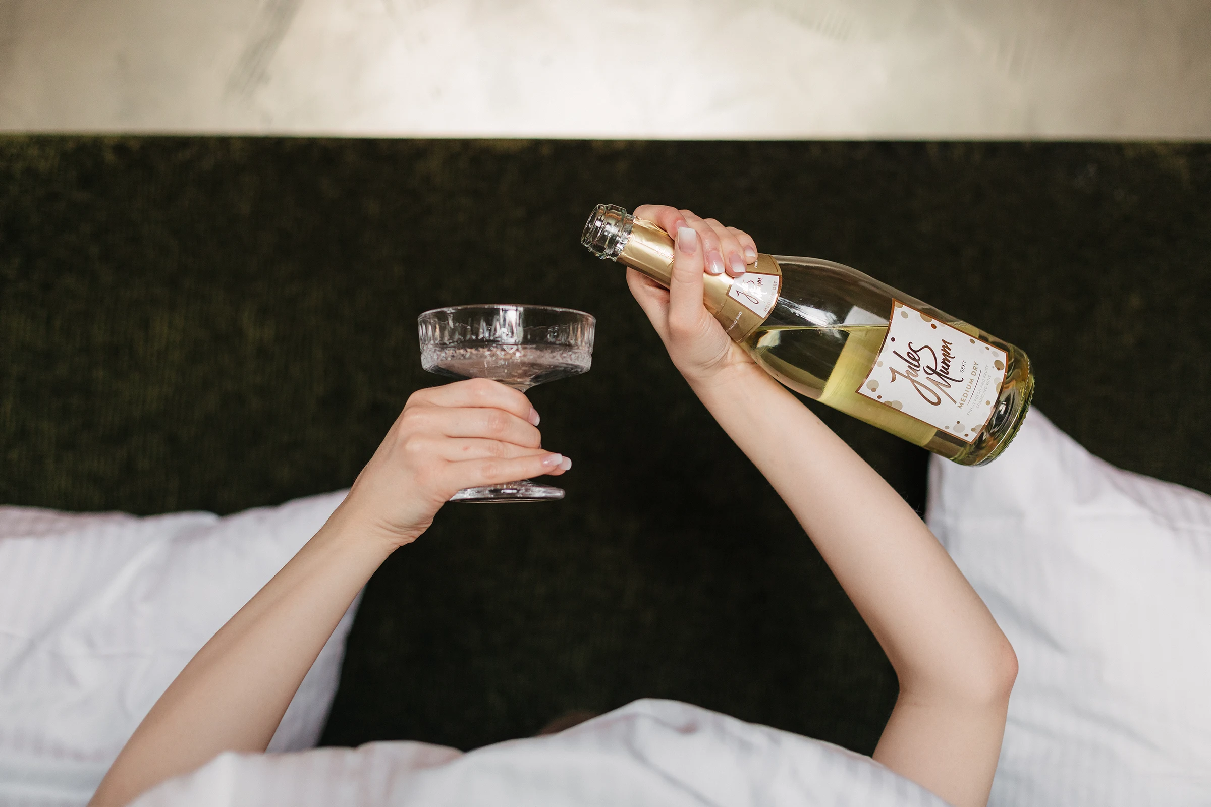 Braut liegt entspannt auf dem Bett und schenkt Sekt in eine Schale ein, lustiges Getting Ready Foto.