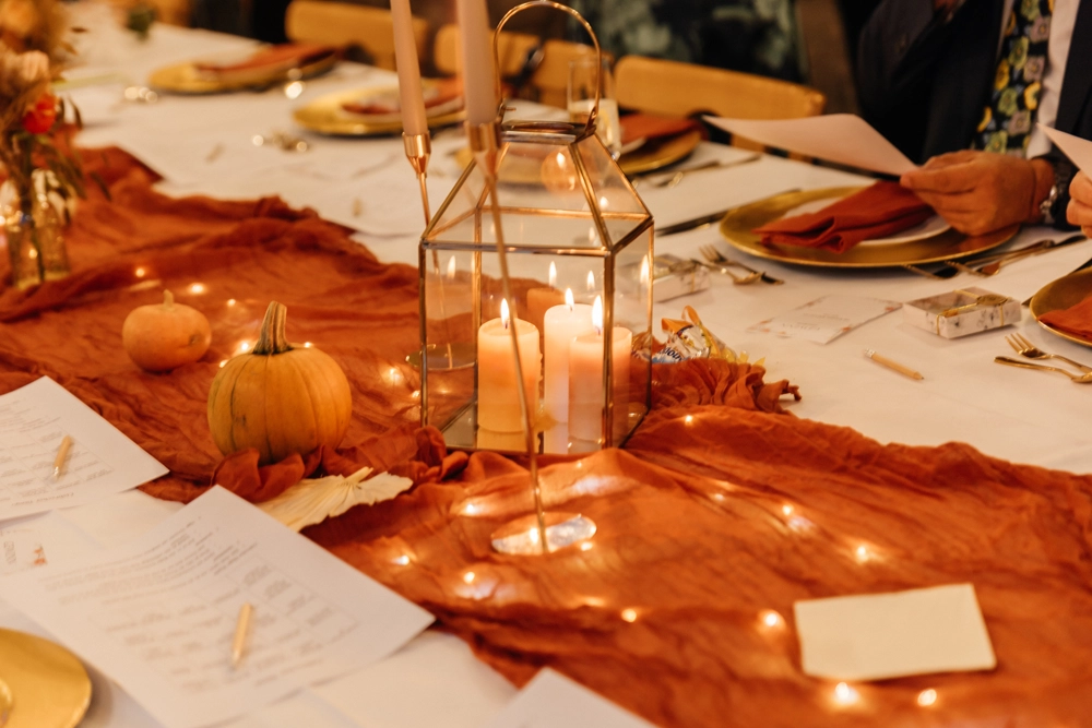 Detaillierte Tischdeko einer Herbsthochzeit: Terrakotta-Tischläufer, kleine Zierkürbisse, goldene Laterne mit Blockkerzen und Lichterketten.