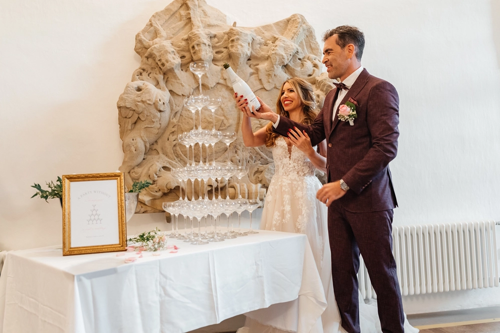 Brautpaar befüllt lachend einen Champagne Tower auf Schloss Hemhofen. Eleganter Sekt-Empfang.