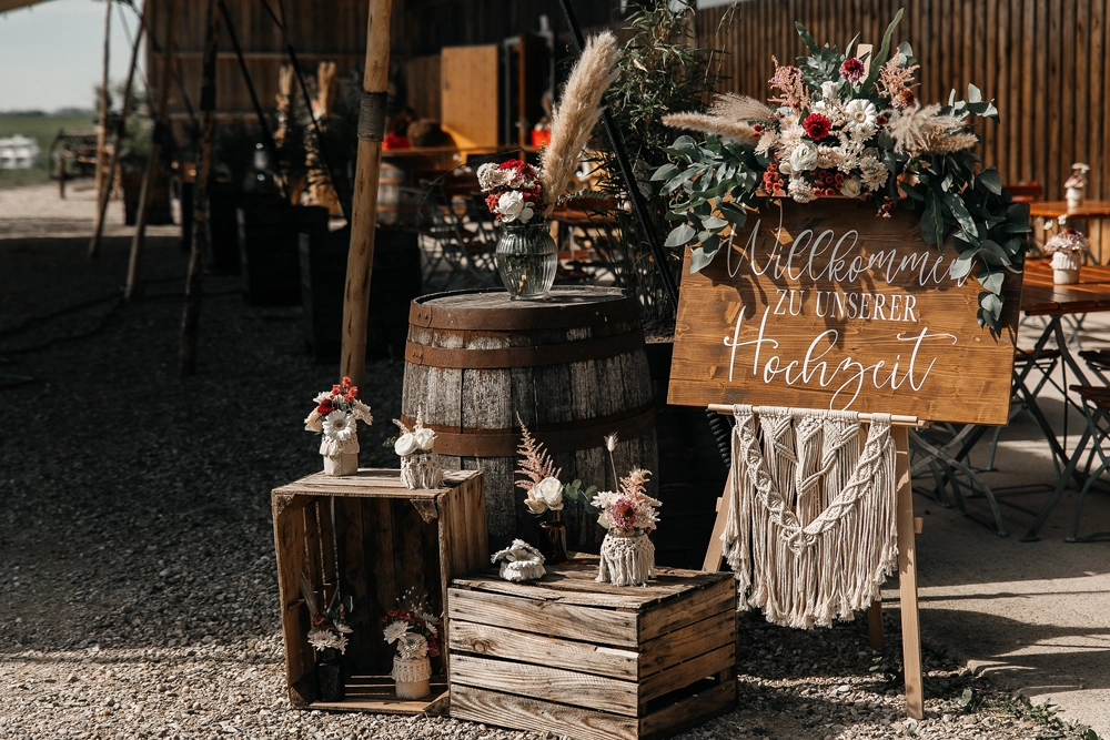 Rustikales Willkommensschild aus Holz mit weißer Kalligrafie und Makramee-Behang auf einer Staffelei. Dekoriert mit Blumenarrangements in Rot und Weiß, Weinfässern und Obstkisten im Vintage-Stil