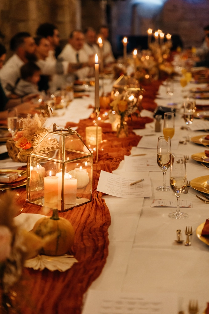 Lange Hochzeitstafel in der Perspektive fotografiert. Der Fokus liegt auf der vorderen Deko (Kerzen, Terrakotta-Läufer), während die sitzenden Gäste im Hintergrund in stimmungsvoller Unschärfe verschwimmen.