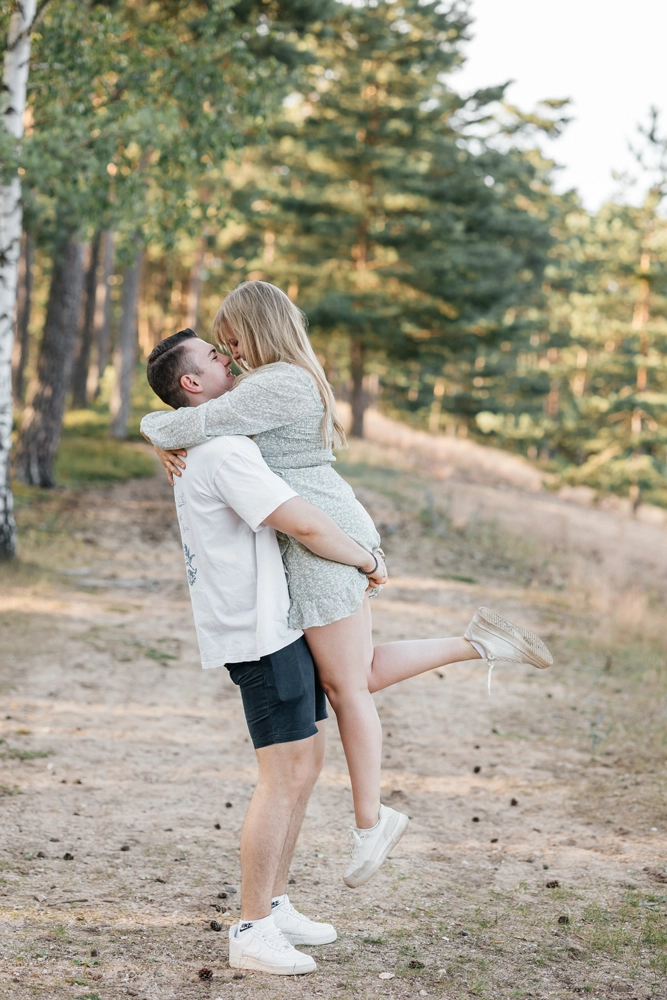 Dynamisches Foto im Wald: Bräutigam hebt die Braut hoch, umgeben von hohen Kiefern.