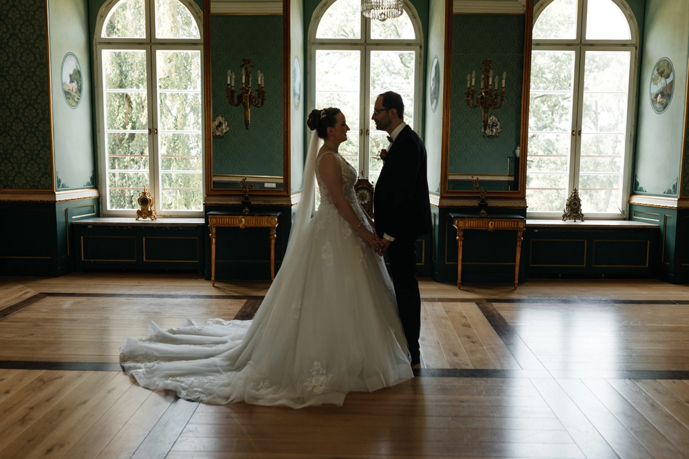 Braut und Bräutigam stehen sich im Grünen Salon von Schloss Jägersburg gegenüber. Das Gegenlicht betont die Silhouette des Paares und die lange Schleppe des Brautkleides auf dem historischen Parkettboden.