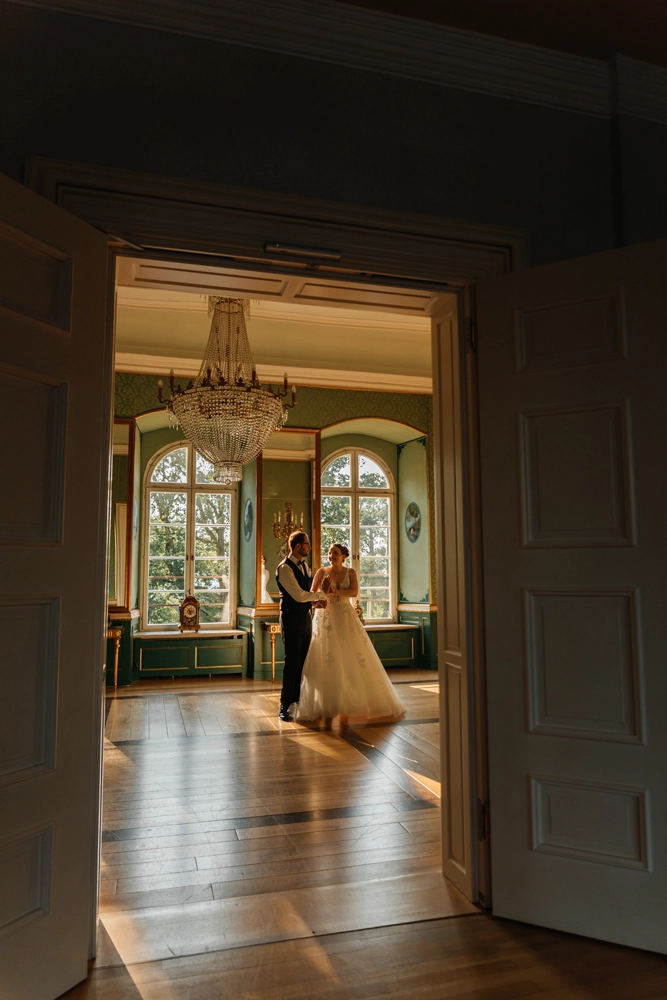 Intimer Moment: Braut und Bräutigam üben ihren ersten Tanz im leeren Festsaal von Schloss Jägersburg. Stimmungsvolles Gegenlicht und historischer Kronleuchter, eingefangen durch die geöffneten Flügeltüren.