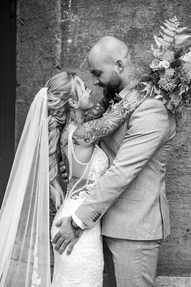 Schwarz-Weiß Hochzeitsfoto: Tätowierte Braut im Spitzenkleid mit langem Schleier umarmt ihren Bräutigam vor einer rustikalen Steinmauer.