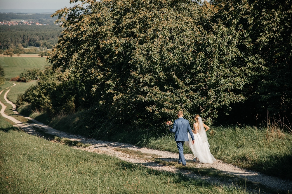 Weitwinkelaufnahme: Brautpaar spaziert Hand in Hand unter großen grünen Bäumen.