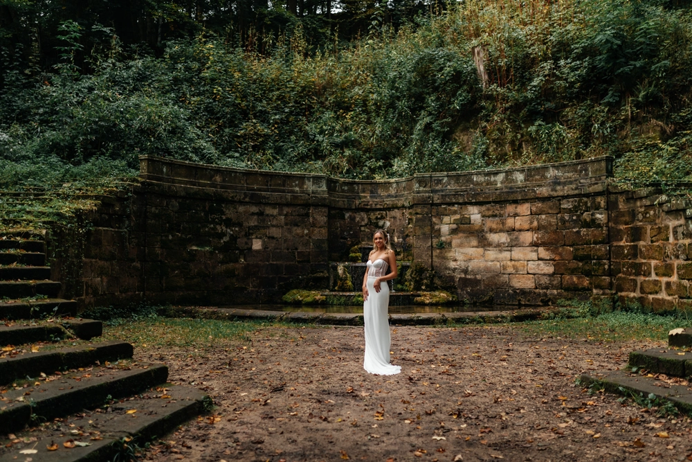 Weitwinkelaufnahme einer Braut in einem modernen, rückenfreien Brautkleid vor einer majestätischen Steinmauer-Kulisse in Nürnberg.