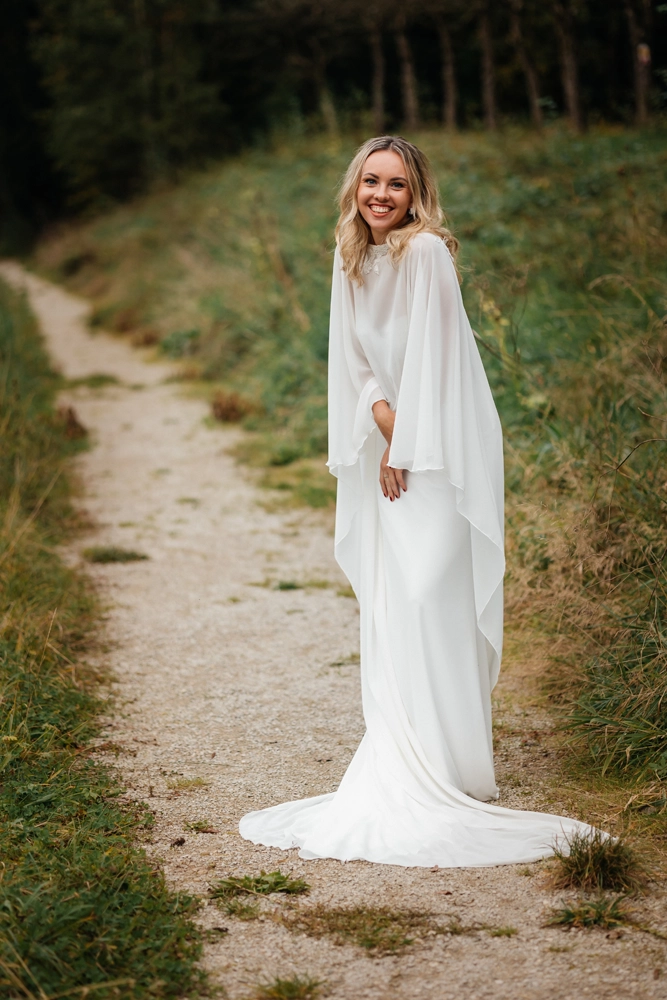 Braut in einem fließenden, transparenten Cape auf einem Kiesweg in natürlicher Umgebung.
