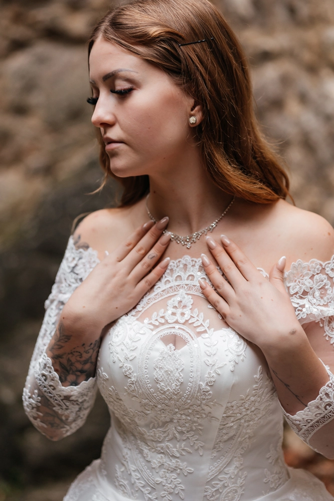 Close-Up auf das Dekolleté der Braut, ihre Hand ruht sanft auf der Brust, Fokus auf den filigranen Halsschmuck und die floralen Spitzendetails.