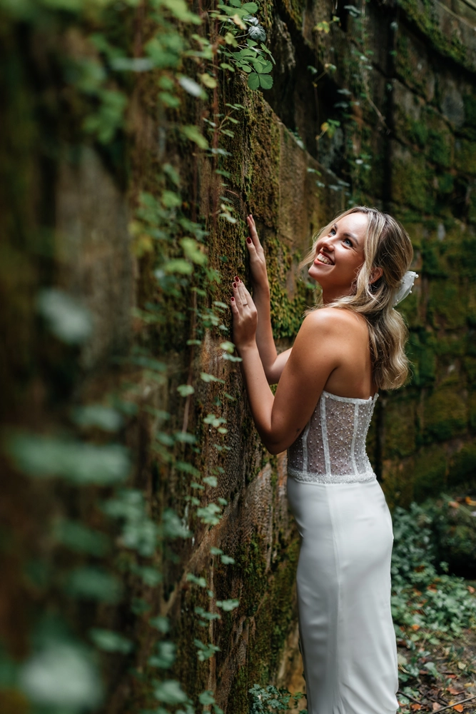 Stimmungsvolles Foto der Braut, die lächelnd an einer mit Efeu bewachsenen Steinwand emporblickt.
