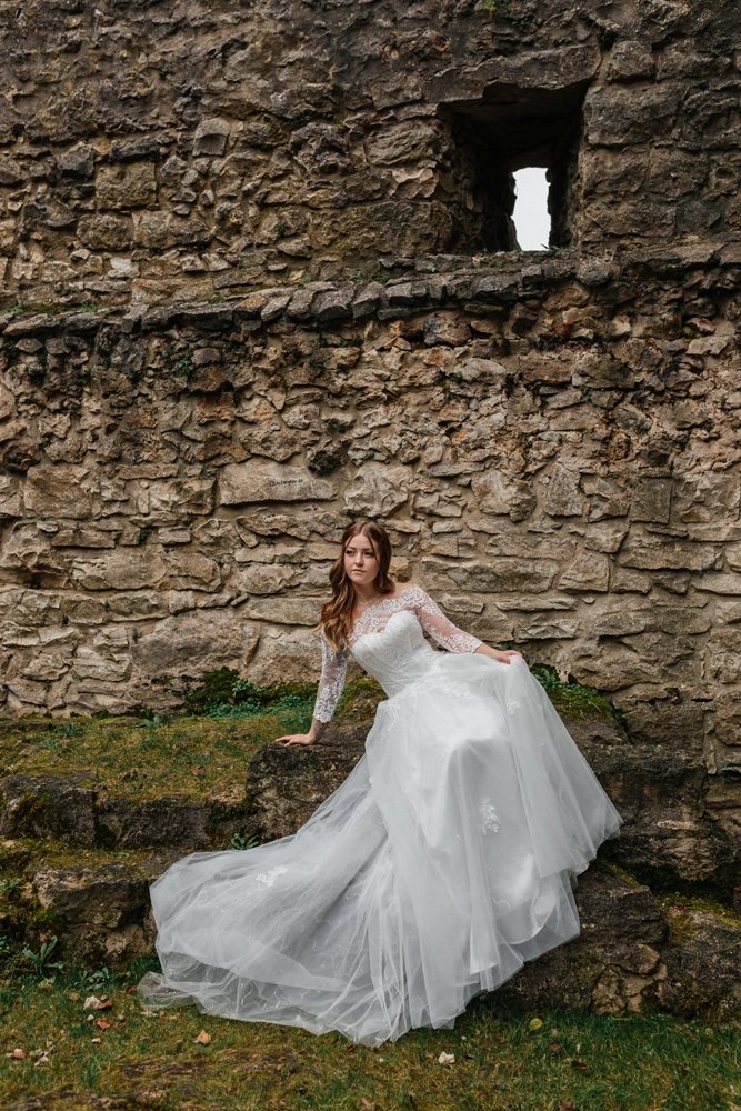Braut sitzt elegant auf den Steinen der Ruine, das weite Tüllkleid ist malerisch um sie herum auf dem Felsen drapiert.