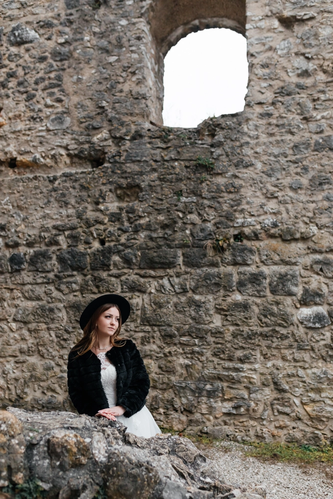 Editorial Brautshooting: Braut sitzt auf Steinen und trägt einen schwarzen Hut sowie einen schwarzen Pelzmantel über dem weißen Brautkleid als modischen Stilbruch.
