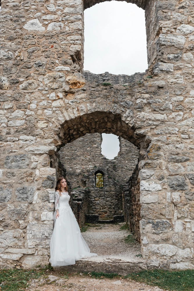 Die Braut steht im weißen Tüllkleid eingerahmt von einem großen, verfallenen Torbogen der Burgruine Wolfstein.