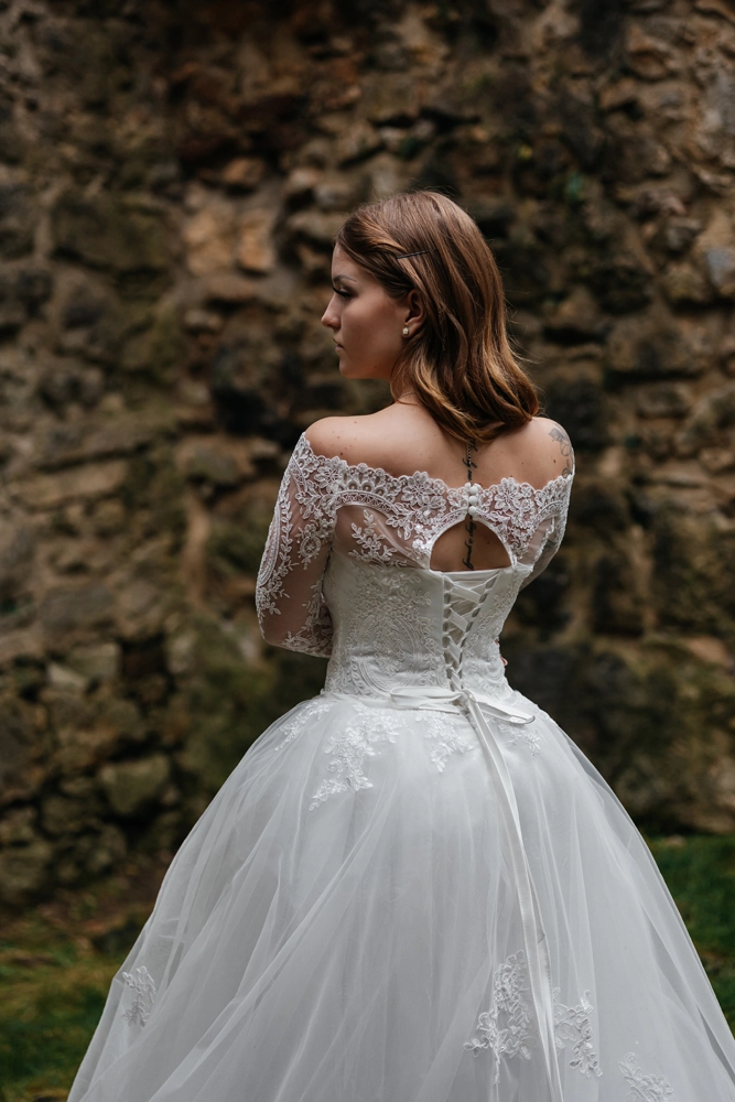 Detailaufnahme der Braut von hinten, die den eleganten Rückenausschnitt aus feiner Spitze des Hochzeitskleides vor einer rustikalen Steinmauer zeigt.