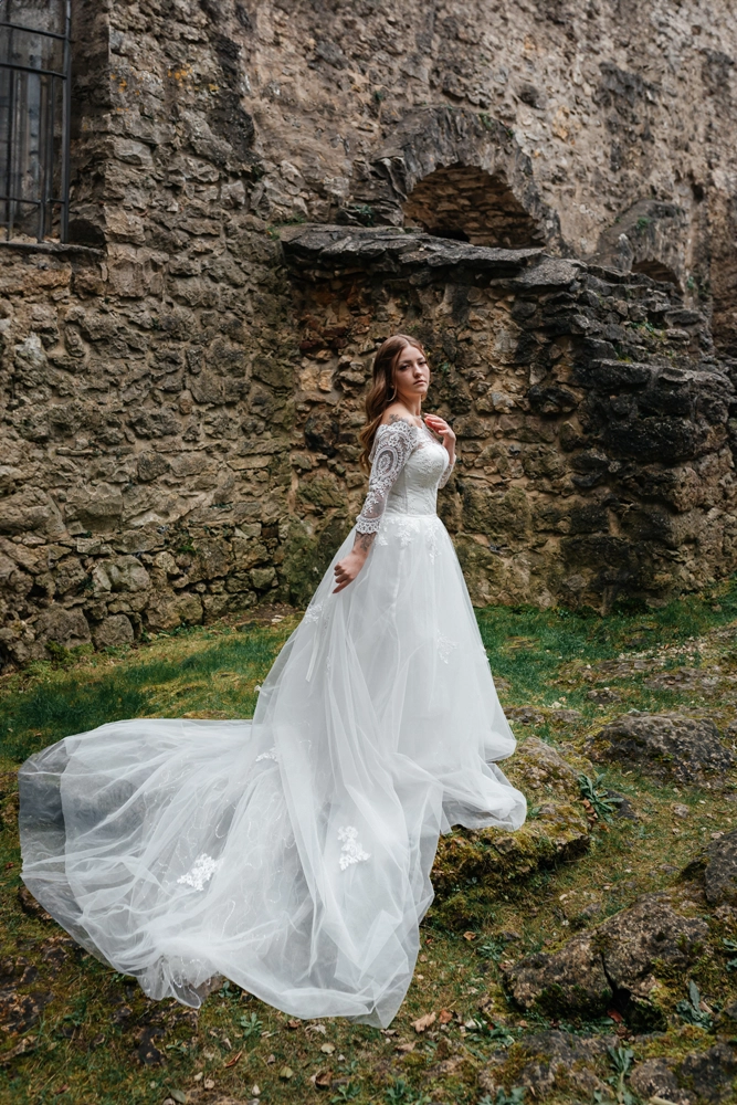 Ganzkörperportrait einer Braut, die mit ausladendem Tüllkleid auf einem moosigen Felsen vor der rauen Steinmauer der Burgruine Wolfstein steht und nach oben blickt.