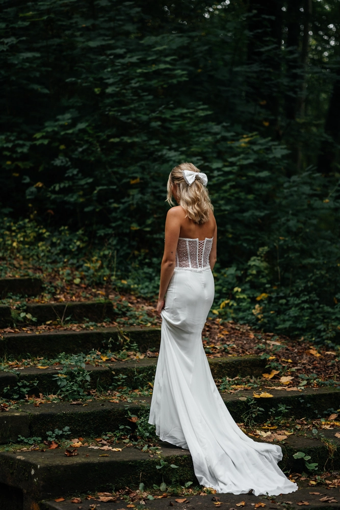 Rückansicht einer Braut auf einer mit Laub bedeckten Waldtreppe, die die lange Schleppe ihres Kleides im Editorial-Stil präsentiert.