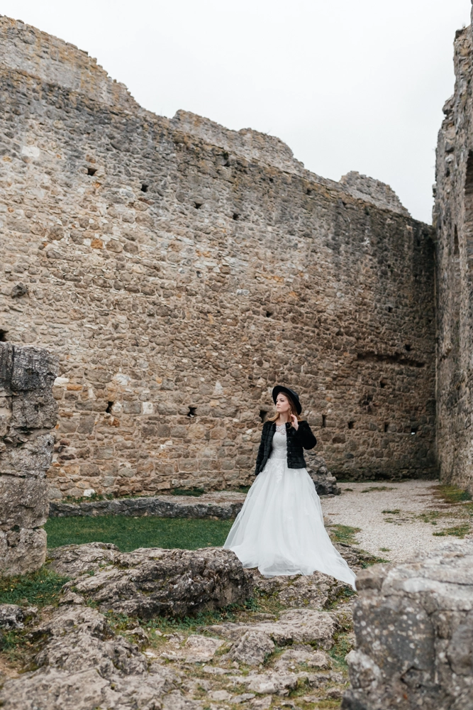 Braut steht im Innenhof der Burgruine Wolfstein, gekleidet in ein weißes Brautkleid kombiniert mit einem markanten schwarzen Pelzmantel und schwarzem Hut.