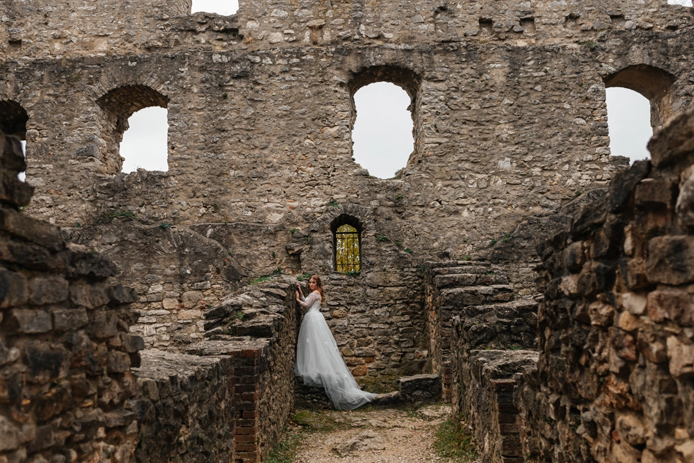 Weitwinkelaufnahme einer Braut im Spitzenkleid mit langer Schleppe, die in der historischen Burgruine Wolfstein durch einen steinernen Torbogen blickt. Dramatischer Nebel im Vordergrund.